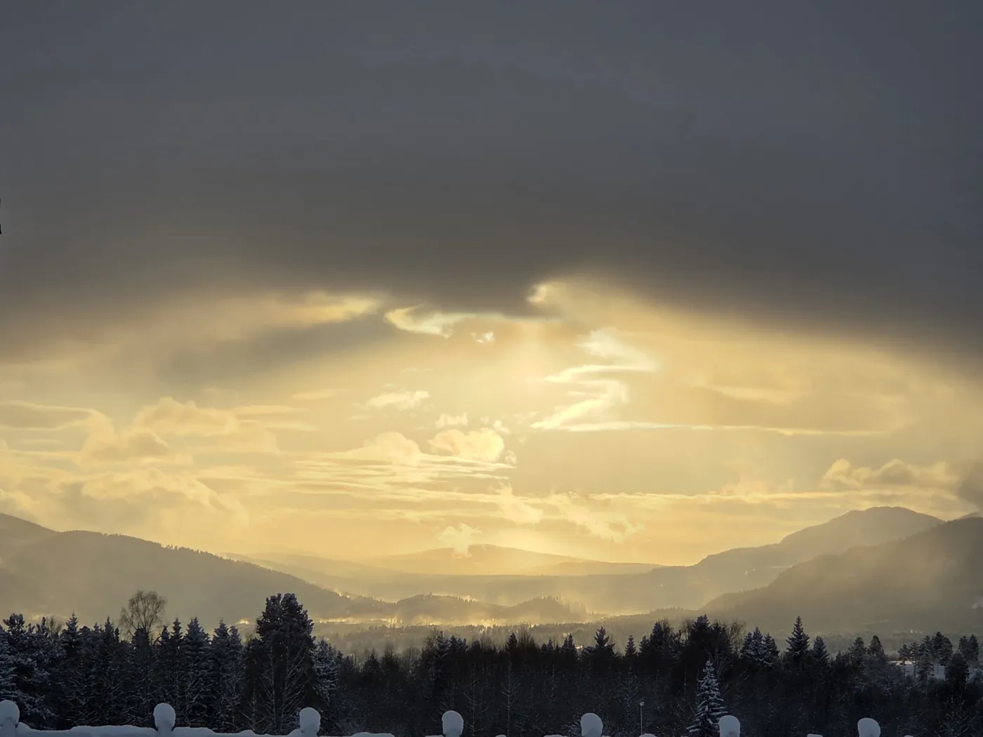 Samme utsikt en vinterkveld: snødekt beite, det hvite gjerdet, fjellene i dalen og en gul solnedgang under tunge skyer